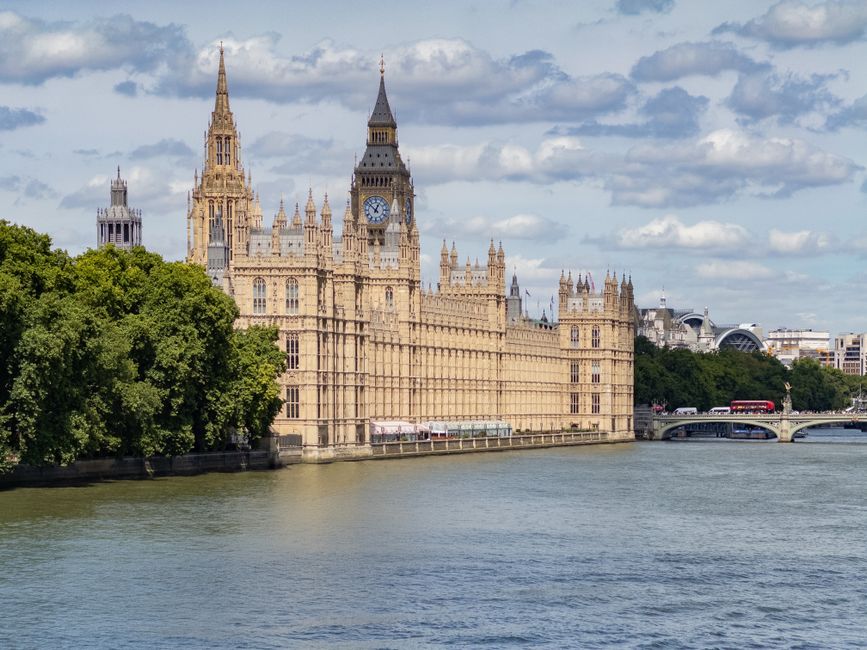A scenic view of the Houses of Parliament and Big Ben in Westminster, London, taken from the River Thames on a partly cloudy day. The historic Gothic-style building features intricate stonework, numer