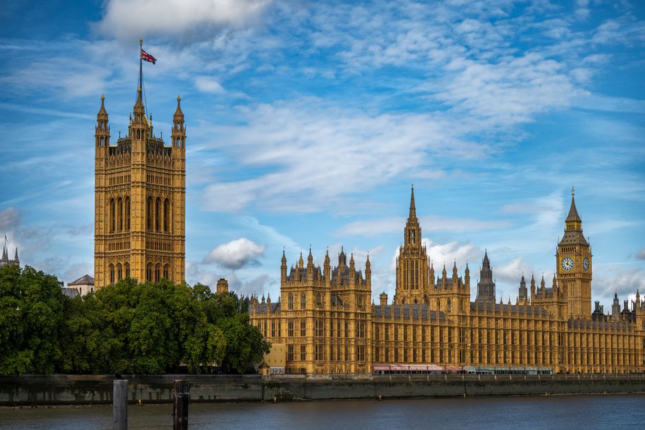 A scenic view of the Houses of Parliament and Big Ben in Westminster, London, taken from the River Thames on a partly cloudy day. The historic Gothic-style building features intricate stonework, numer